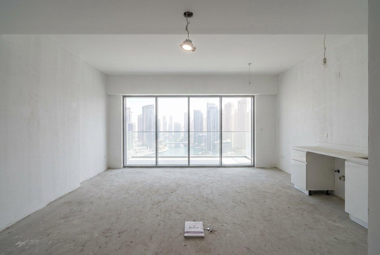 Bare-shell Dubai Marina apartment at developer handover, raw cement floor, unfinished walls, floor-to-ceiling window with Marina view